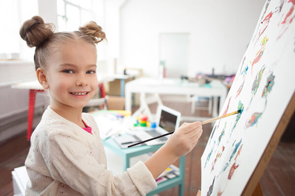 Pretty small painter is creating a picture on the canvas. The girl is standing and smiling. She is looking at camera happily
